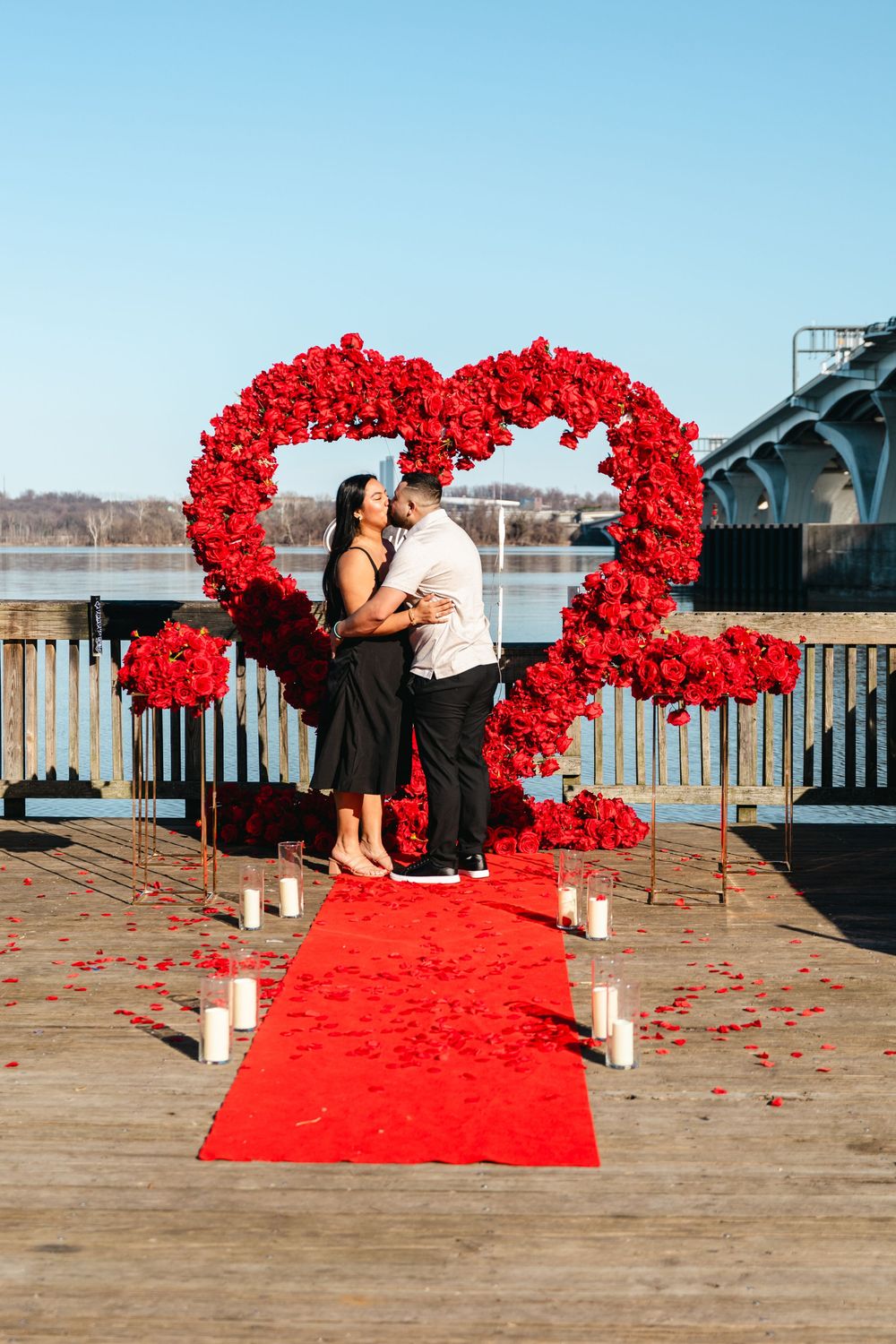 Couple kissing on a red carpet pier, framed by a large red rose heart arch with candles and rose petals.