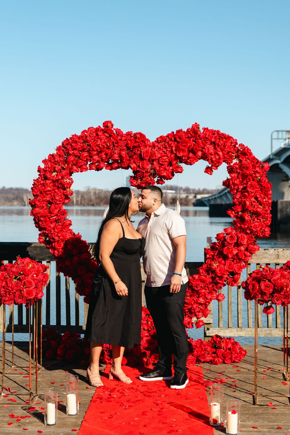 Couple kisses in front of giant red rose heart arch on waterfront dock with candles and rose petals.