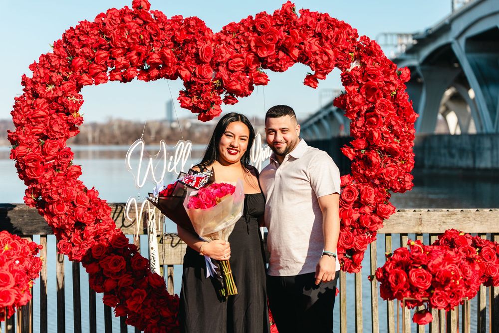 Couple poses in front of large red rose heart frame during romantic marriage proposal by a river bridge.