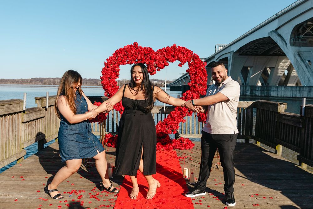 Three people celebrate a romantic proposal near a red rose heart arch on a waterfront dock on a sunny day.