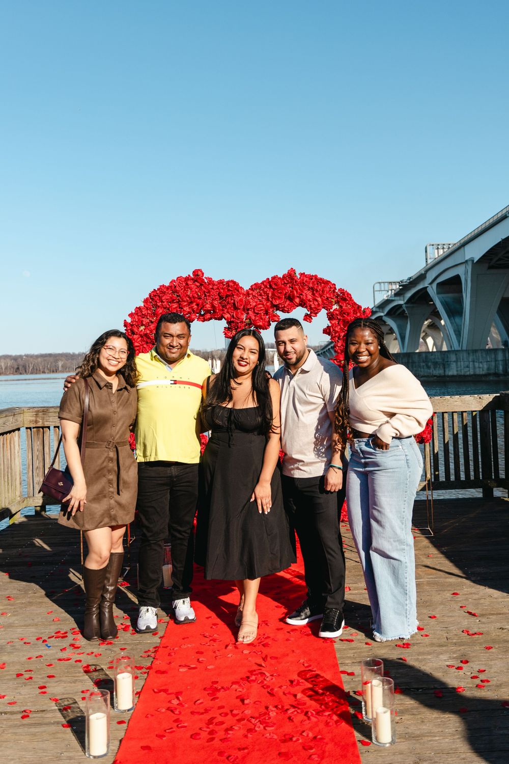 Five friends pose on a red carpet under a floral heart arch on a waterfront dock with candles and rose petals.