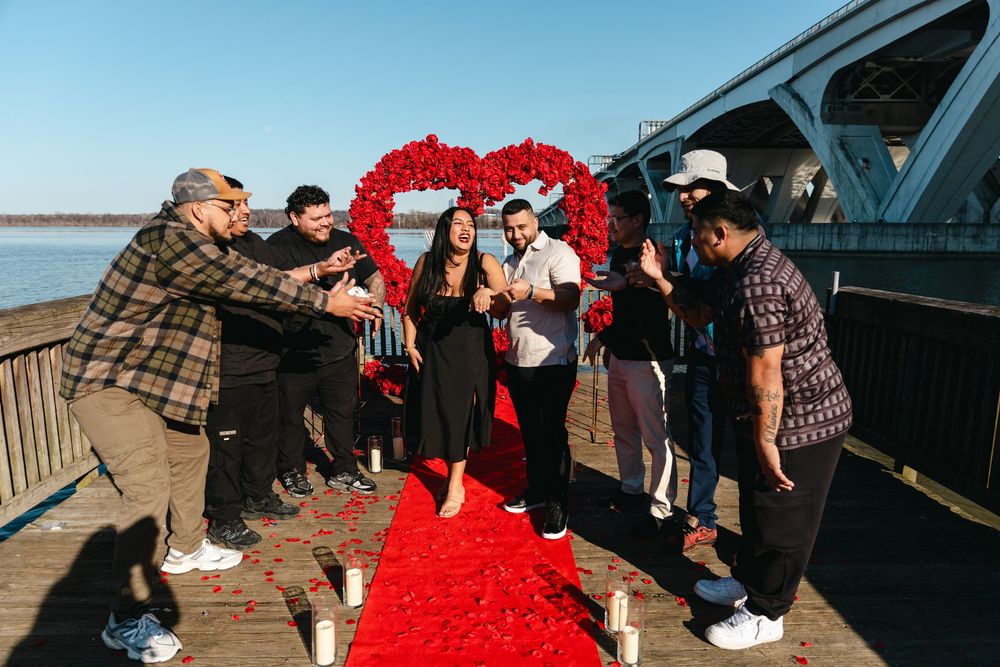 Group celebrating a romantic proposal on a waterfront pier with red rose heart decorations and a red carpet.
