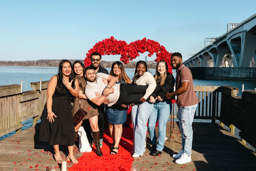 Group of friends celebrating a romantic proposal on a waterfront deck with red roses heart backdrop.