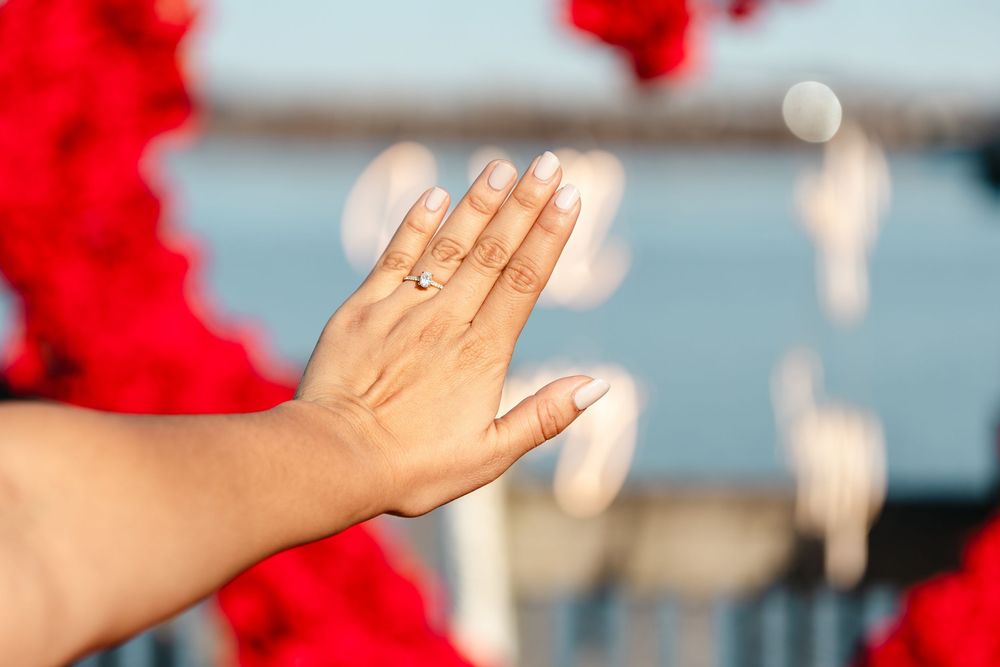 Close-up of a hand displaying an engagement ring with red roses and water in the background.