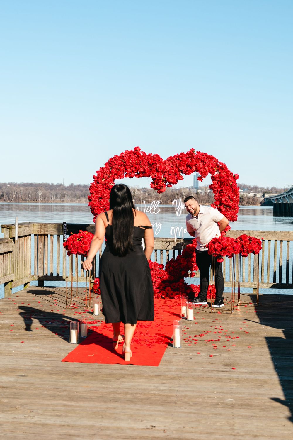 Man surprises woman with romantic marriage proposal on waterfront dock with large red rose heart arch and red carpet.