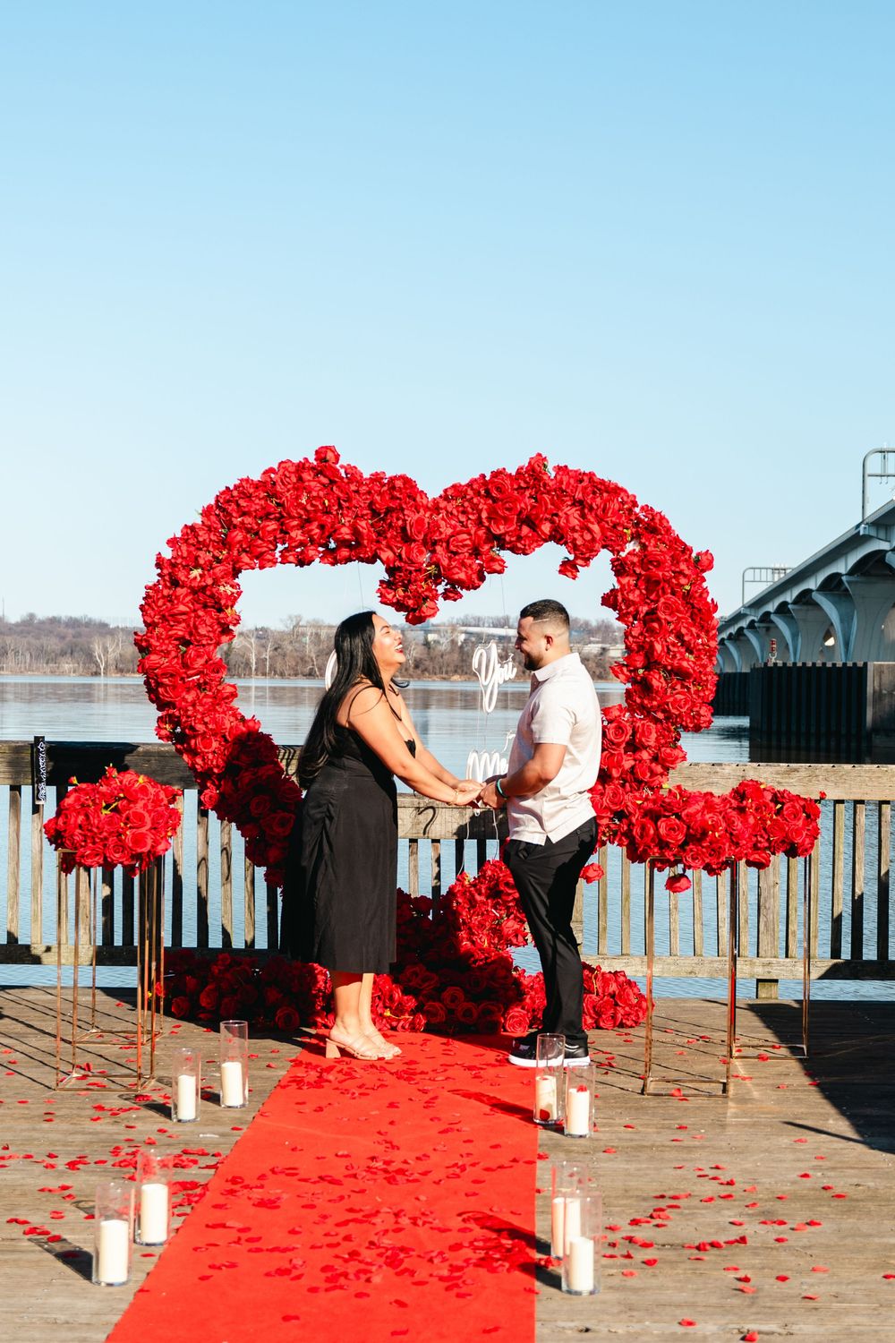 Couple standing under a large red rose heart arch on a waterfront dock with red carpet and candles.