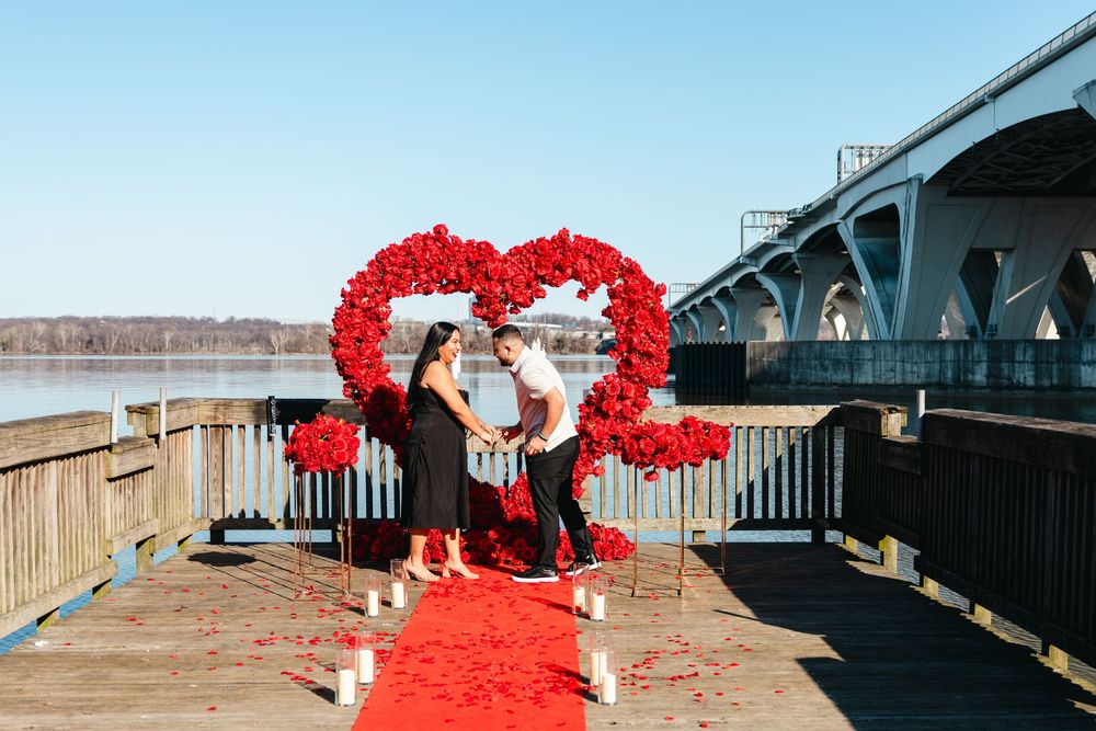 Couple shares romantic proposal moment on waterfront dock with red rose heart arch and red carpet by a bridge.