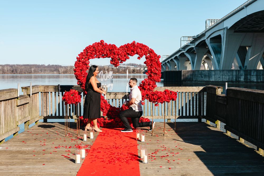 Man proposes to woman on a dock with a red rose heart arch, red carpet, and candles by a river bridge.