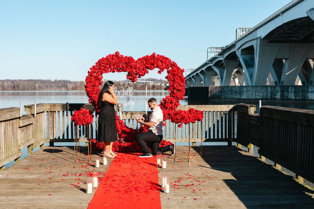Man proposes on a decorated dock with a red rose heart arch, red carpet, and candles by a river bridge.
