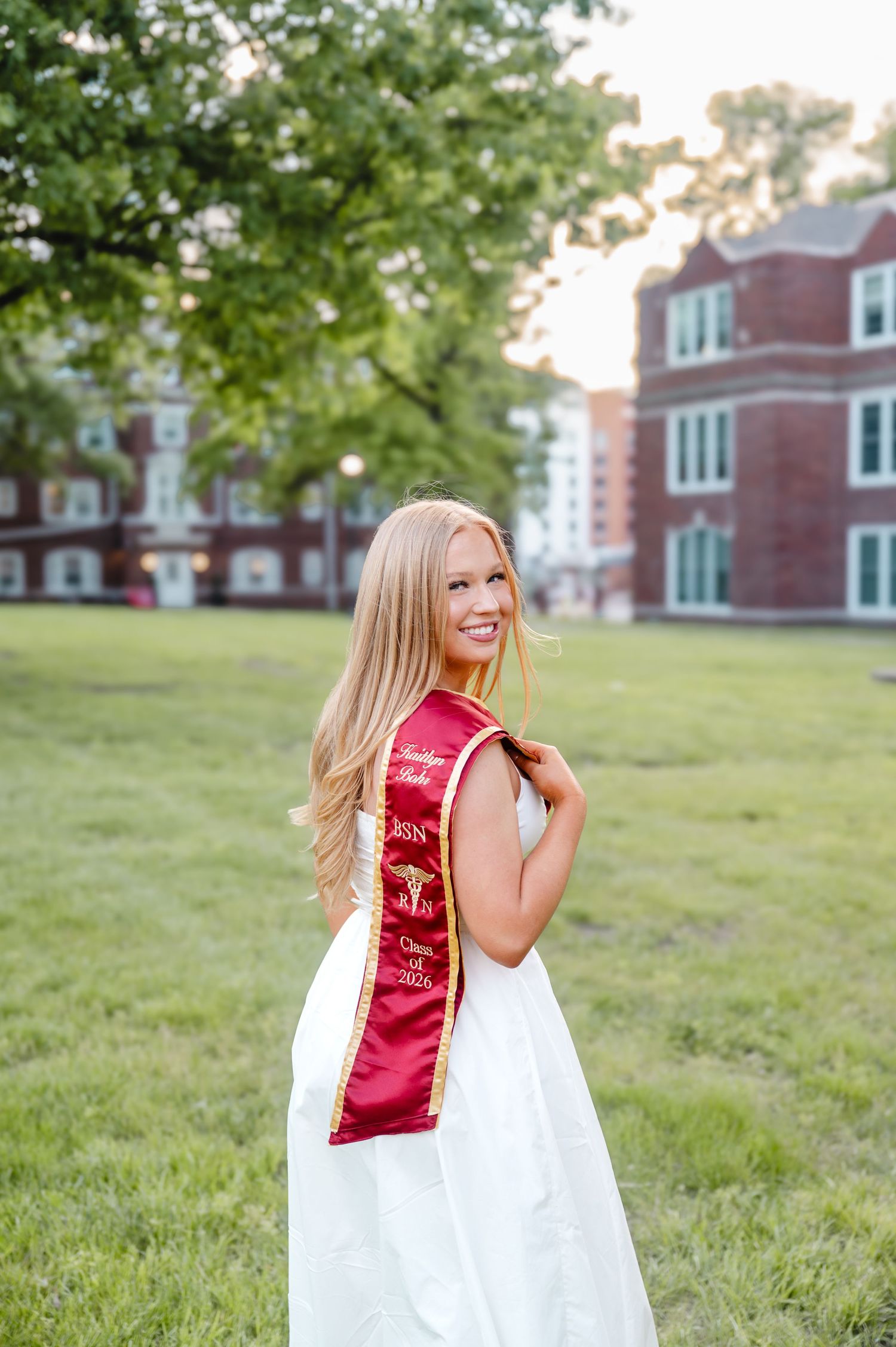 Smiling blonde graduate in white dress and red graduation sash poses outdoors on green campus lawn near brick building.