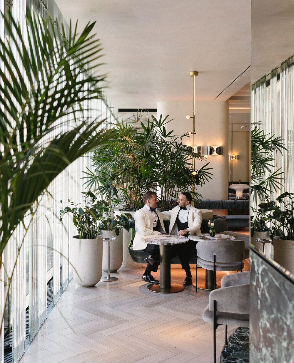 Two elegantly dressed staff members consult at a table in a lush, plant-filled modern restaurant interior.