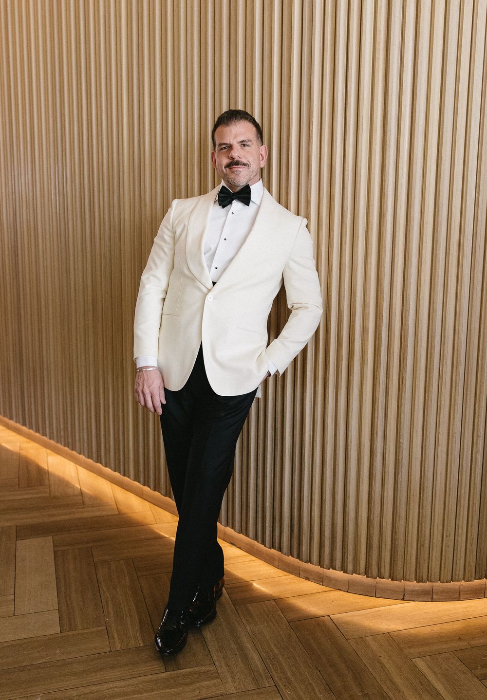 Man in white tuxedo jacket, black bow tie and trousers poses confidently in front of wooden slat wall.
