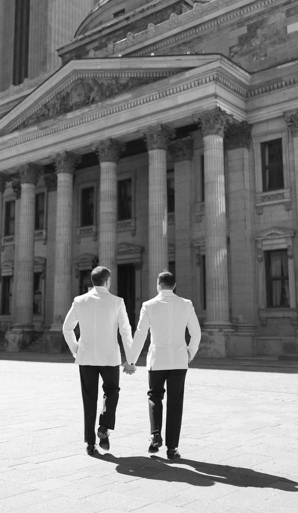 Two men in white suits hold hands walking toward a grand neoclassical building with columns in black and white photo.