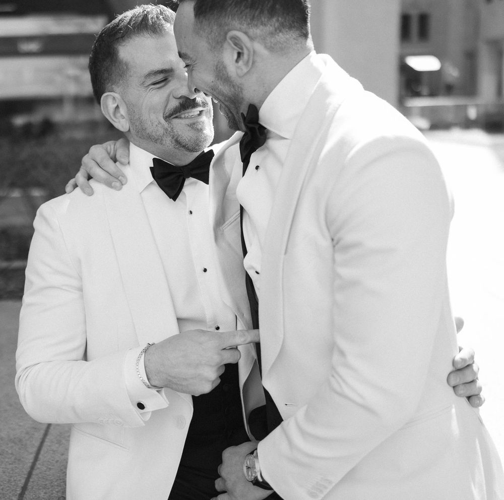 Two grooms in white tuxedos share an intimate moment, smiling and embracing on their wedding day.