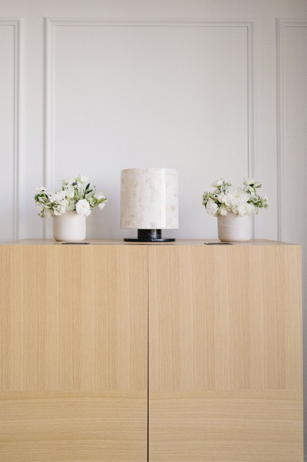 Minimalist wooden sideboard with marble table lamp and white floral arrangements against white paneled wall.