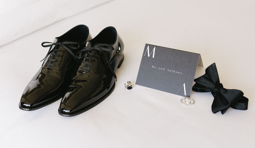 Black patent leather dress shoes with lace-ups, a gray card, black bow tie, and ring on white surface.