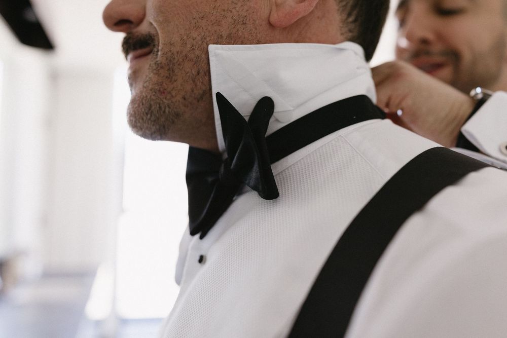 Man in white dress shirt with black bow tie and suspenders being helped by another person getting ready.