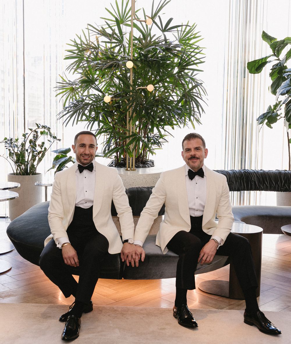 Two grooms in white tuxedo jackets and black bow ties sit together on a curved sofa surrounded by tropical plants.