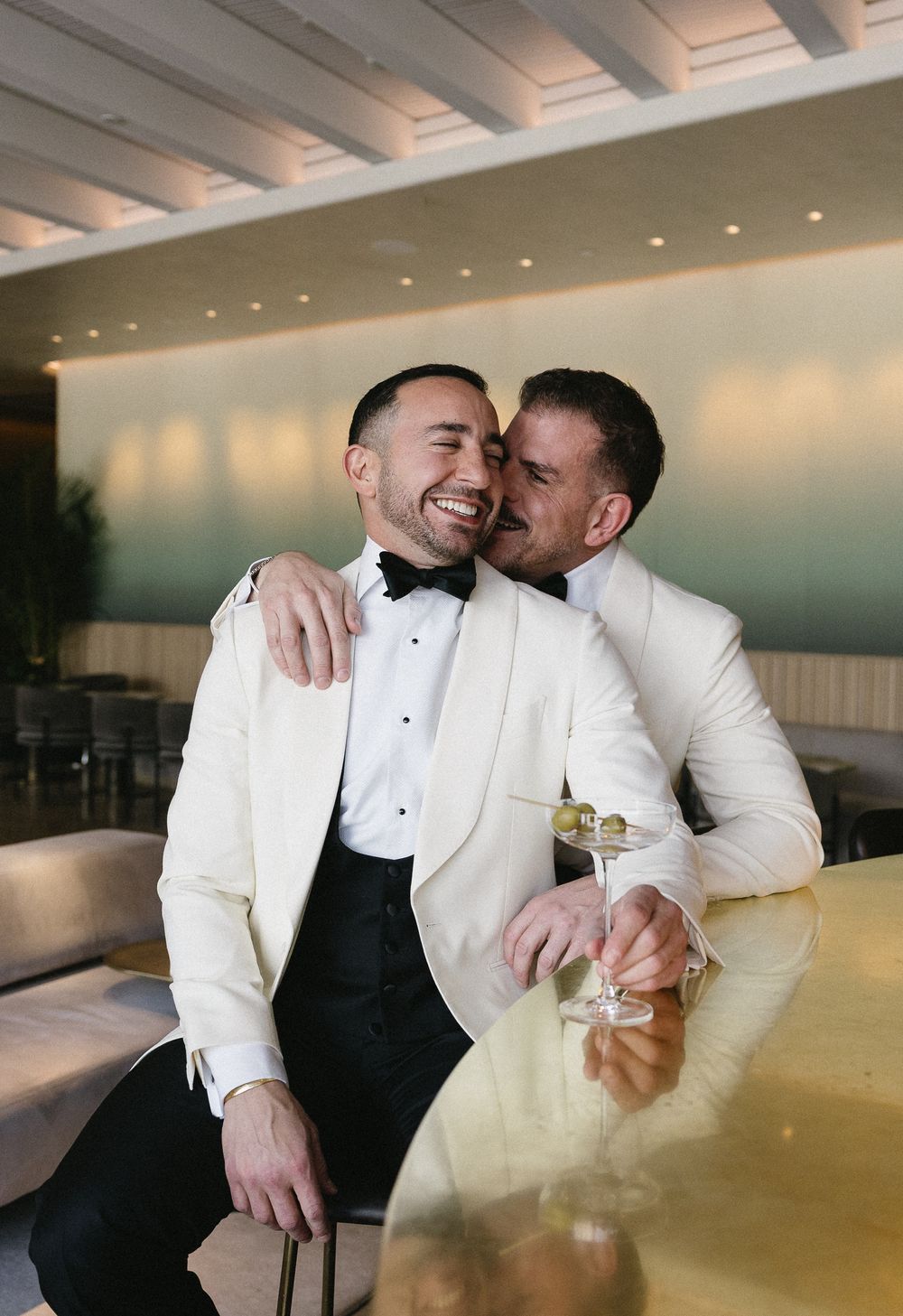 Two grooms in white tuxedo jackets share a joyful embrace at a gold bar, holding champagne glasses at their wedding reception.