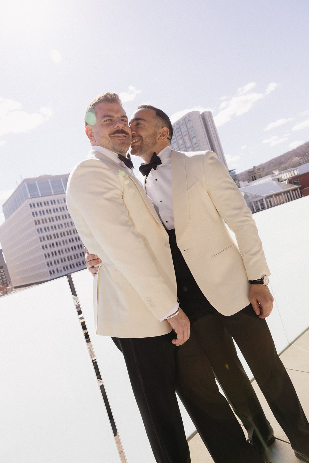 Two grooms in white tuxedo jackets share a tender moment on a rooftop with city skyline backdrop.