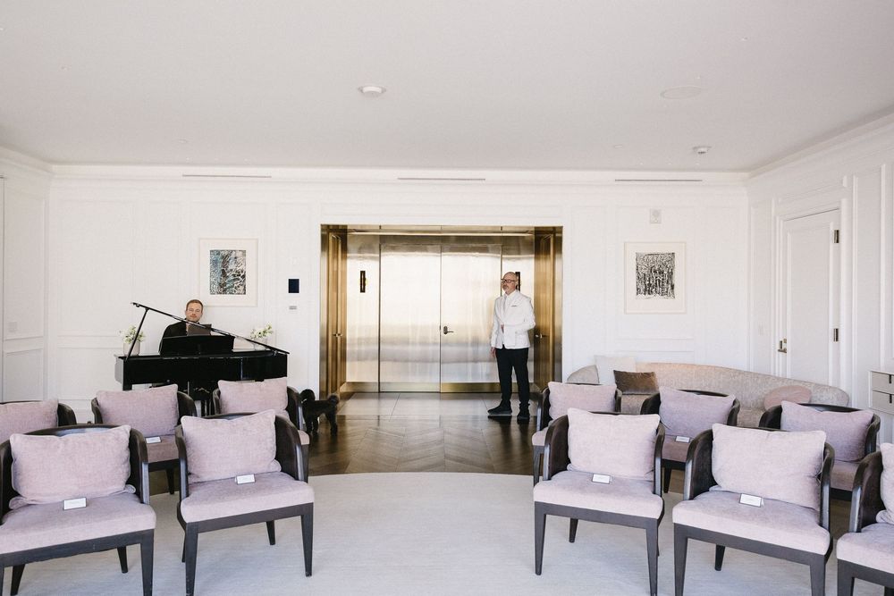 Elegant waiting room with pink chairs, grand piano, and gold elevator doors in a white interior space.