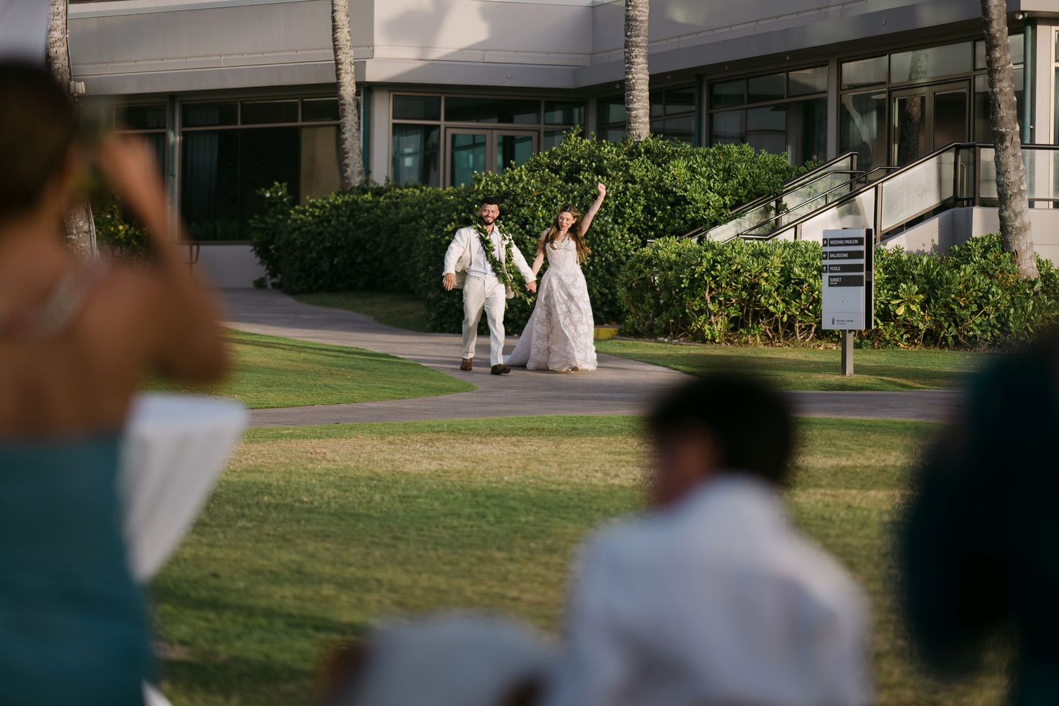 Bride and groom celebrate outdoors on lush green lawn at tropical wedding venue with palm trees.