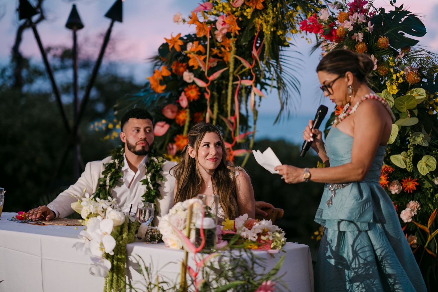 Couple sits at wedding reception table with tropical floral arch backdrop as woman in blue dress gives speech.