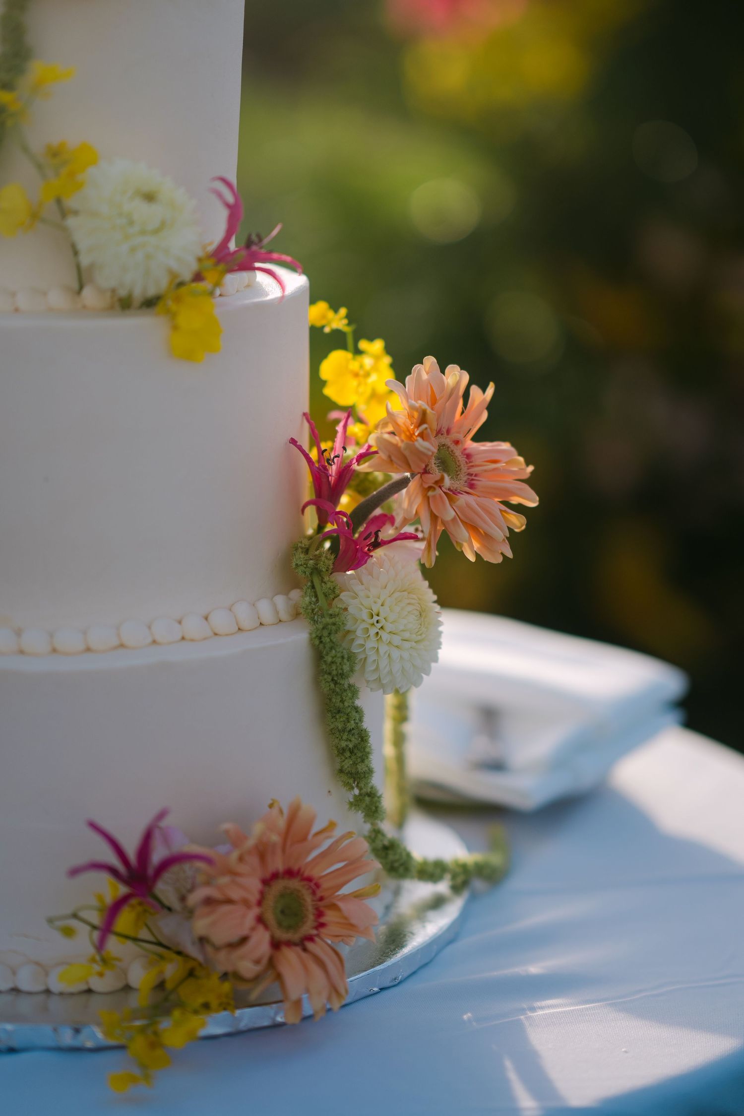 Elegant three-tier white wedding cake adorned with colorful fresh flowers including pink gerbera daisies and yellow blooms outdoors.