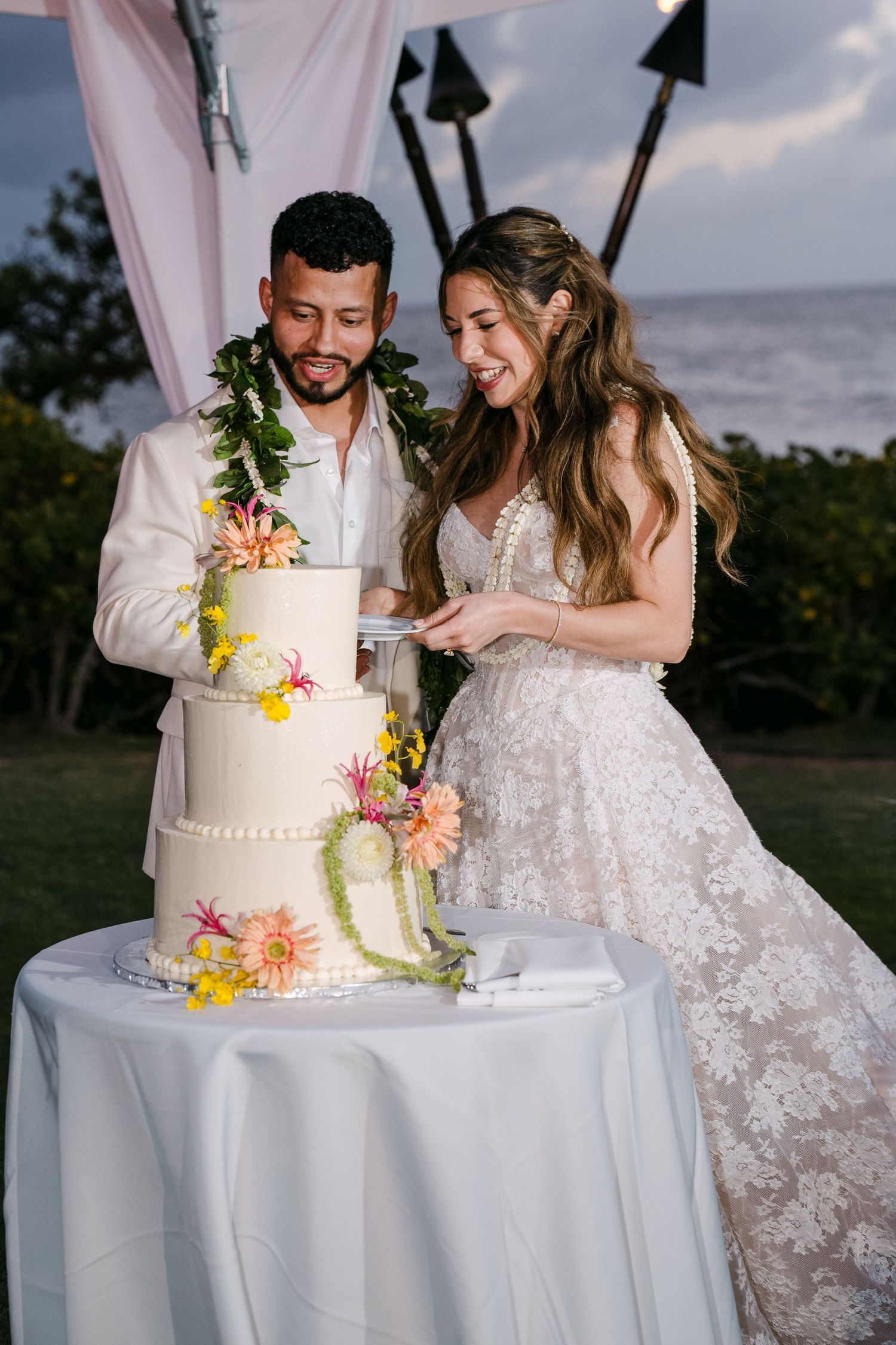 Bride and groom cutting a floral three-tier wedding cake outdoors by the ocean at sunset.
