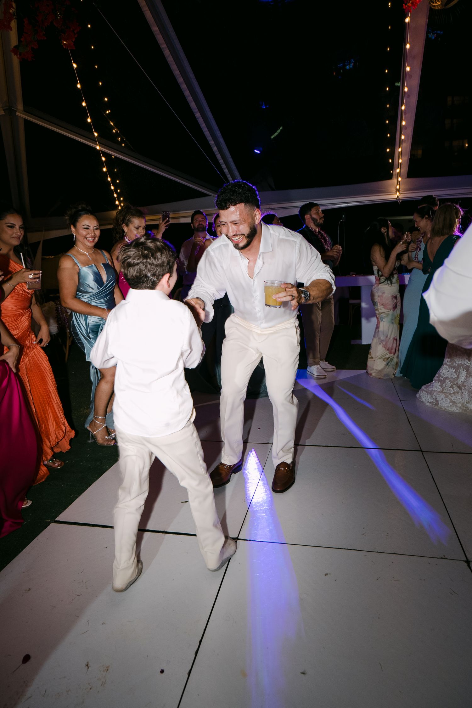 A man and boy in white outfits dance joyfully at an elegant outdoor wedding reception at night.