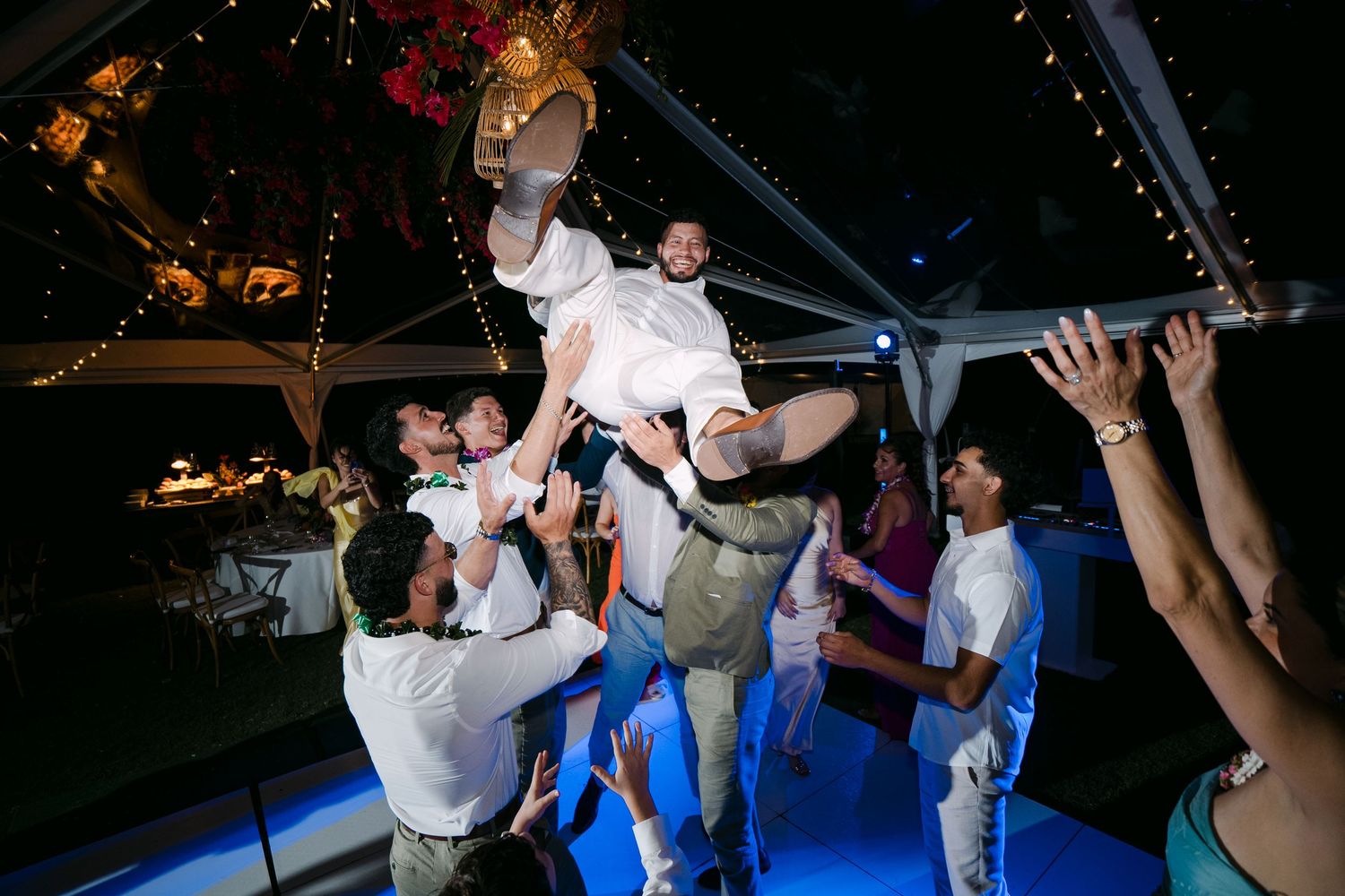 Wedding guests lifting bride and groom on dance floor during lively outdoor evening reception celebration.