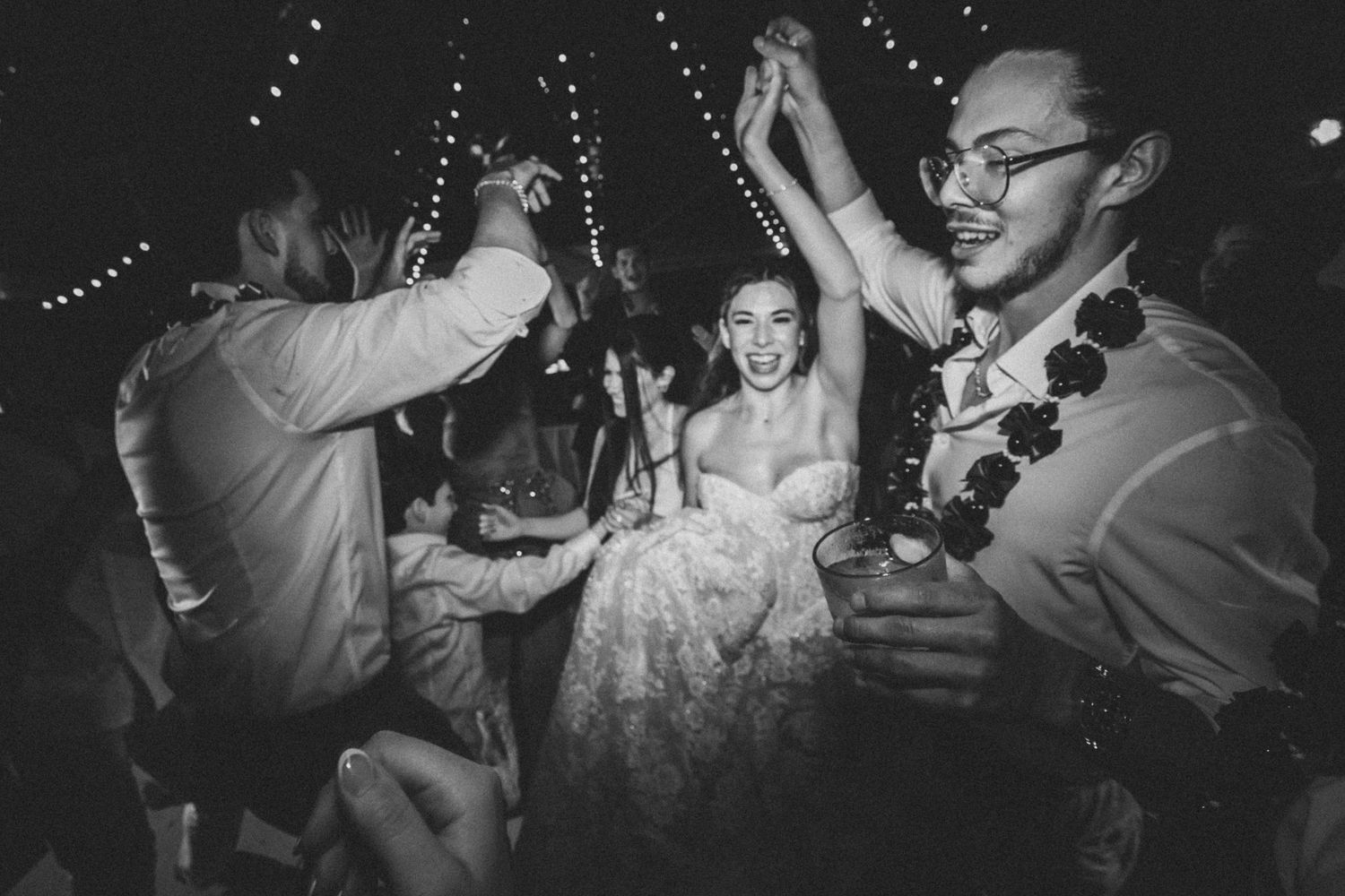 Black and white wedding dance floor photo of bride and groom celebrating joyfully with guests under string lights.