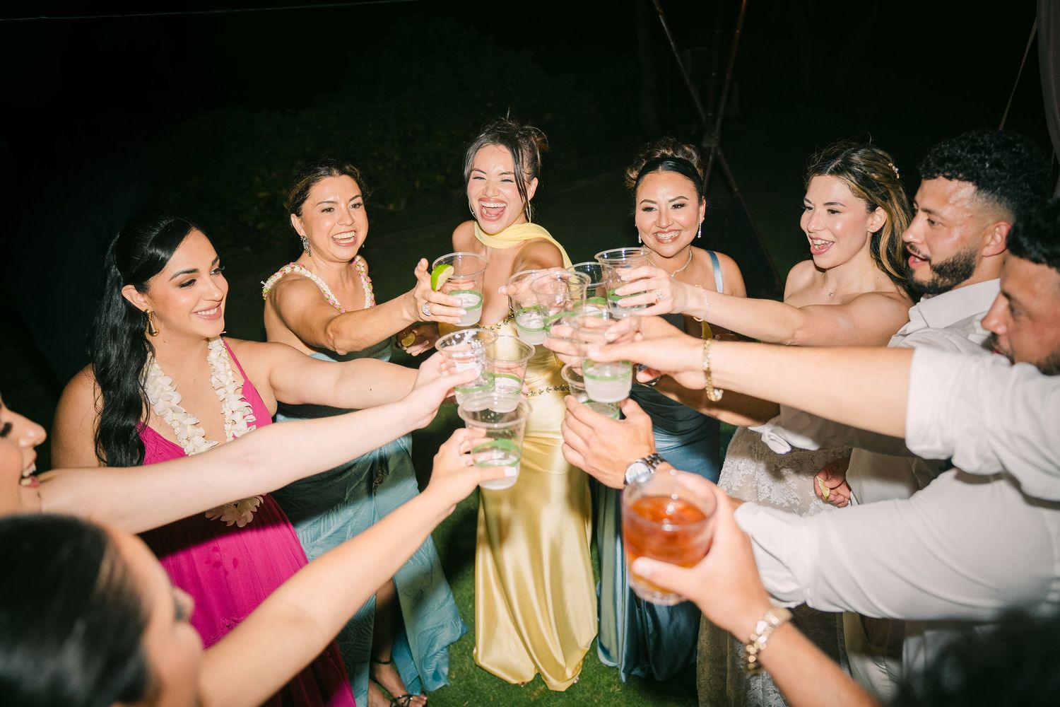 Wedding party guests in formal attire toasting with drinks at a nighttime outdoor celebration.