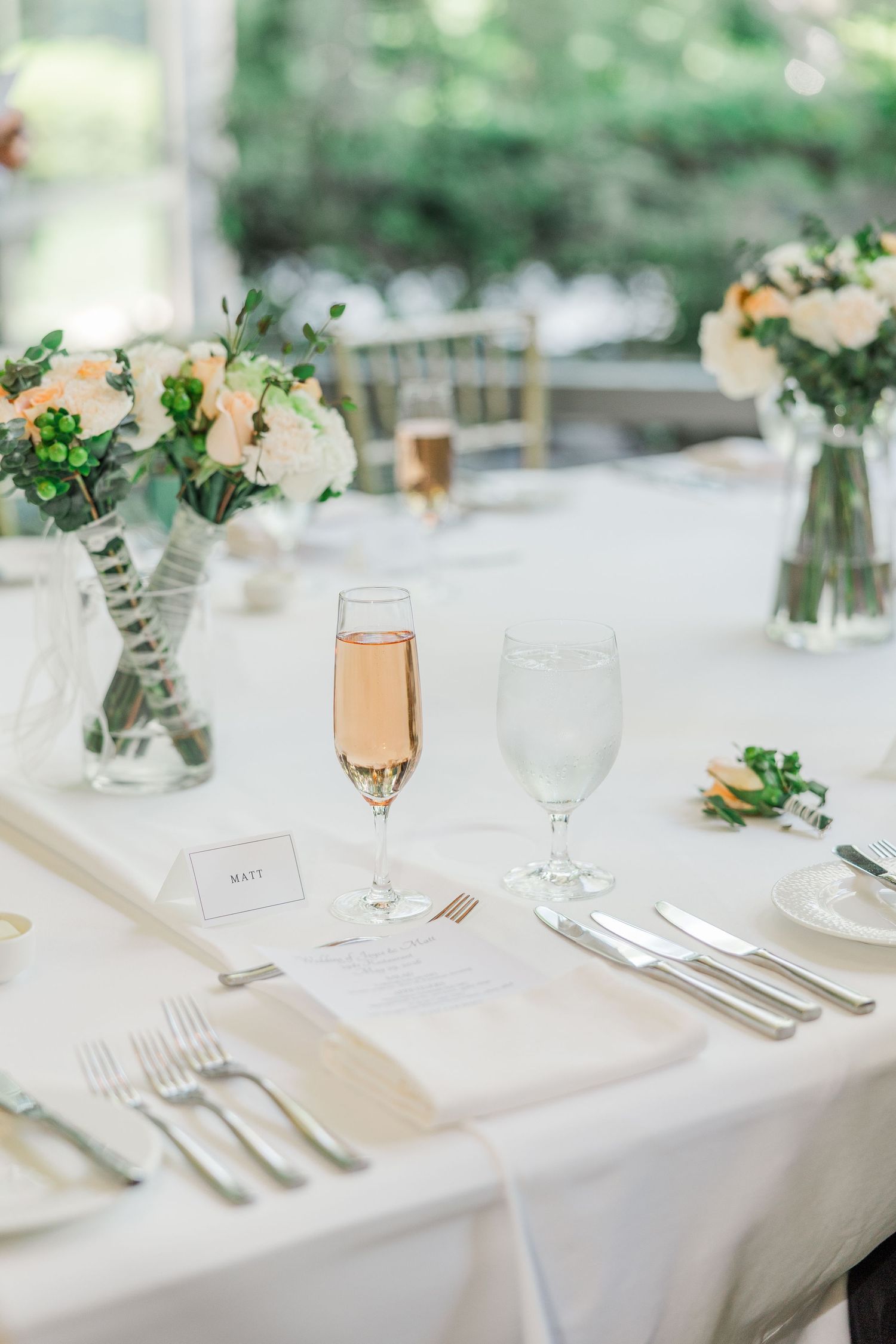 Elegant wedding reception table setting with rose champagne, white florals, place cards, and silverware on a white linen tablecloth.