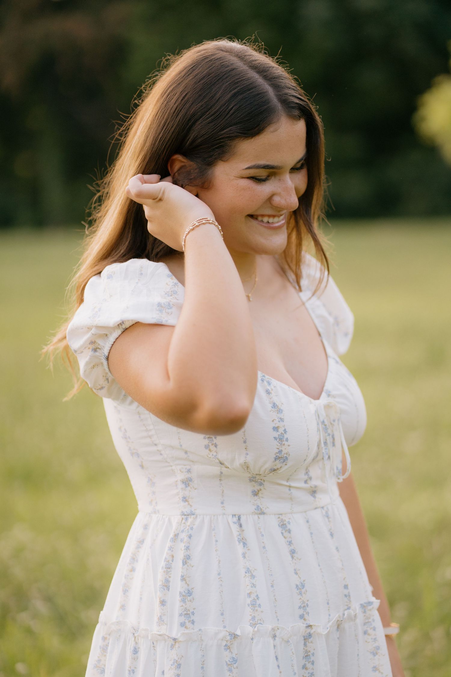 Young woman in white floral dress smiling outdoors in golden sunlight, brushing hair back with hand.