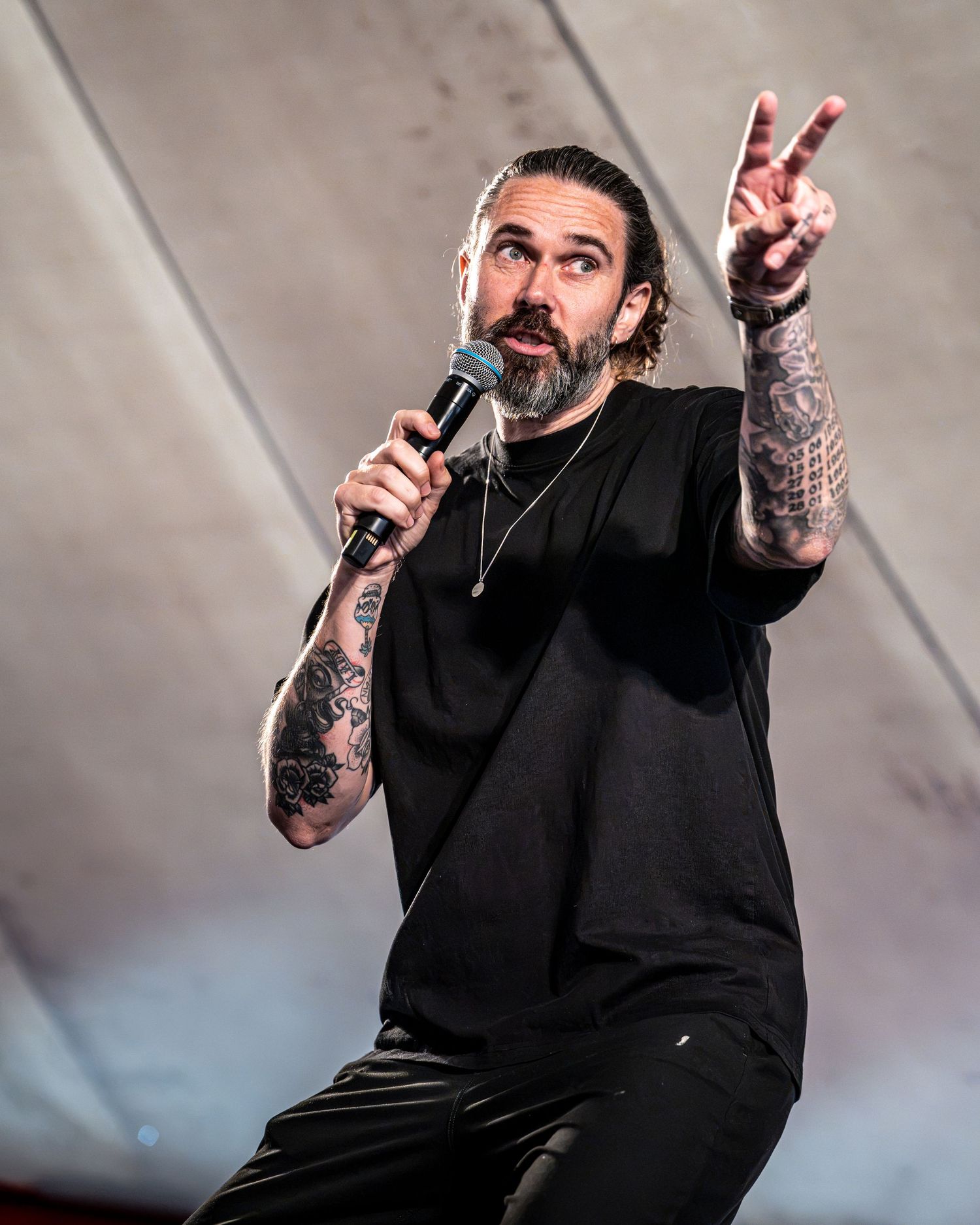 Tattooed male performer singing into microphone on stage, wearing black outfit, gesturing with hand raised.