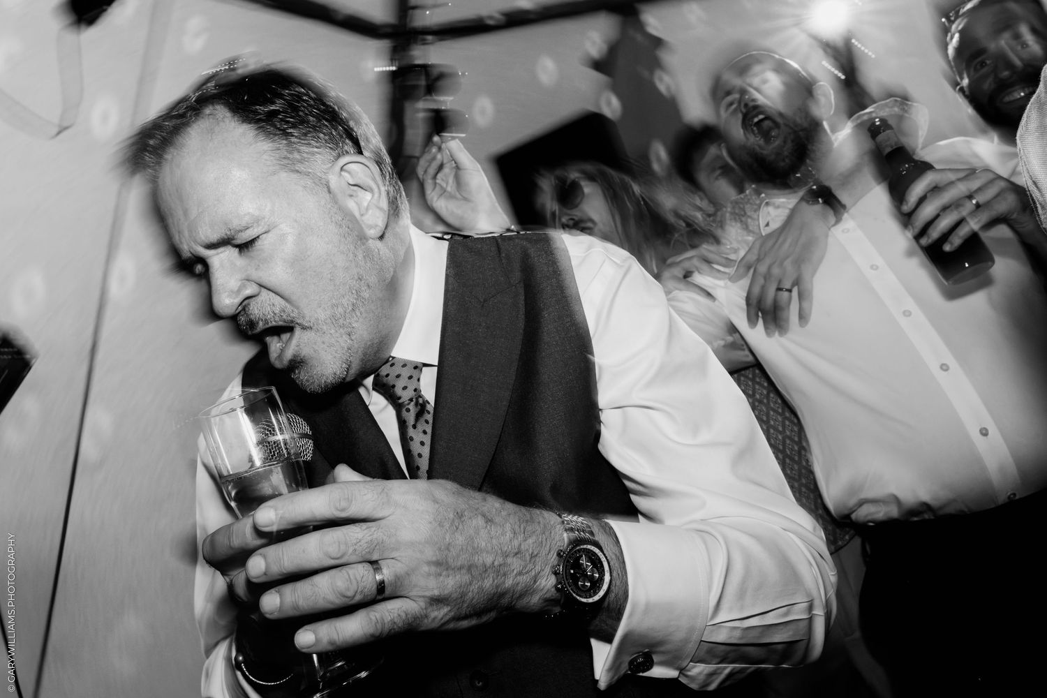 Black and white photo of a man in a vest dancing energetically at a wedding reception holding a drink.
