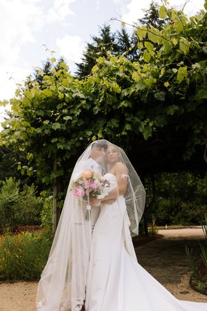 Two brides sharing a romantic kiss under a veil in a lush garden setting.