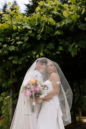 Bride and groom sharing a romantic moment under a floral wedding veil, holding a colorful bouquet outdoors.