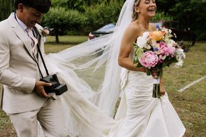 Laughing bride in white gown holds colorful bouquet while groom with camera adjusts her flowing veil outdoors.