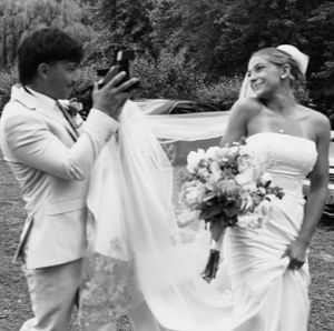 Black and white photo of a bride holding a bouquet while a man in a suit photographs her at an outdoor wedding.