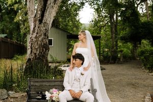 Bride in white gown and veil stands behind groom in ivory suit on bench, surrounded by lush greenery.