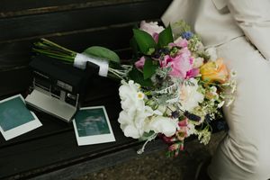 Colorful wedding bouquet with pink, white, and orange flowers beside a Polaroid camera on a wooden bench.