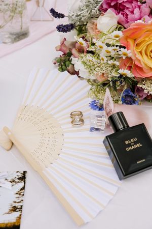 Wedding flat lay featuring a Bleu de Chanel perfume bottle, rings, fan, and colorful floral bouquet.