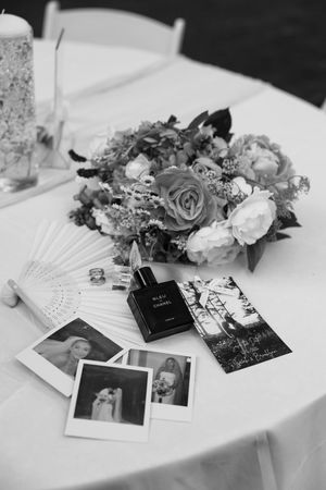 Black and white wedding flat lay with floral bouquet, Polaroid photos, Chanel perfume, fan, and ring on white table.