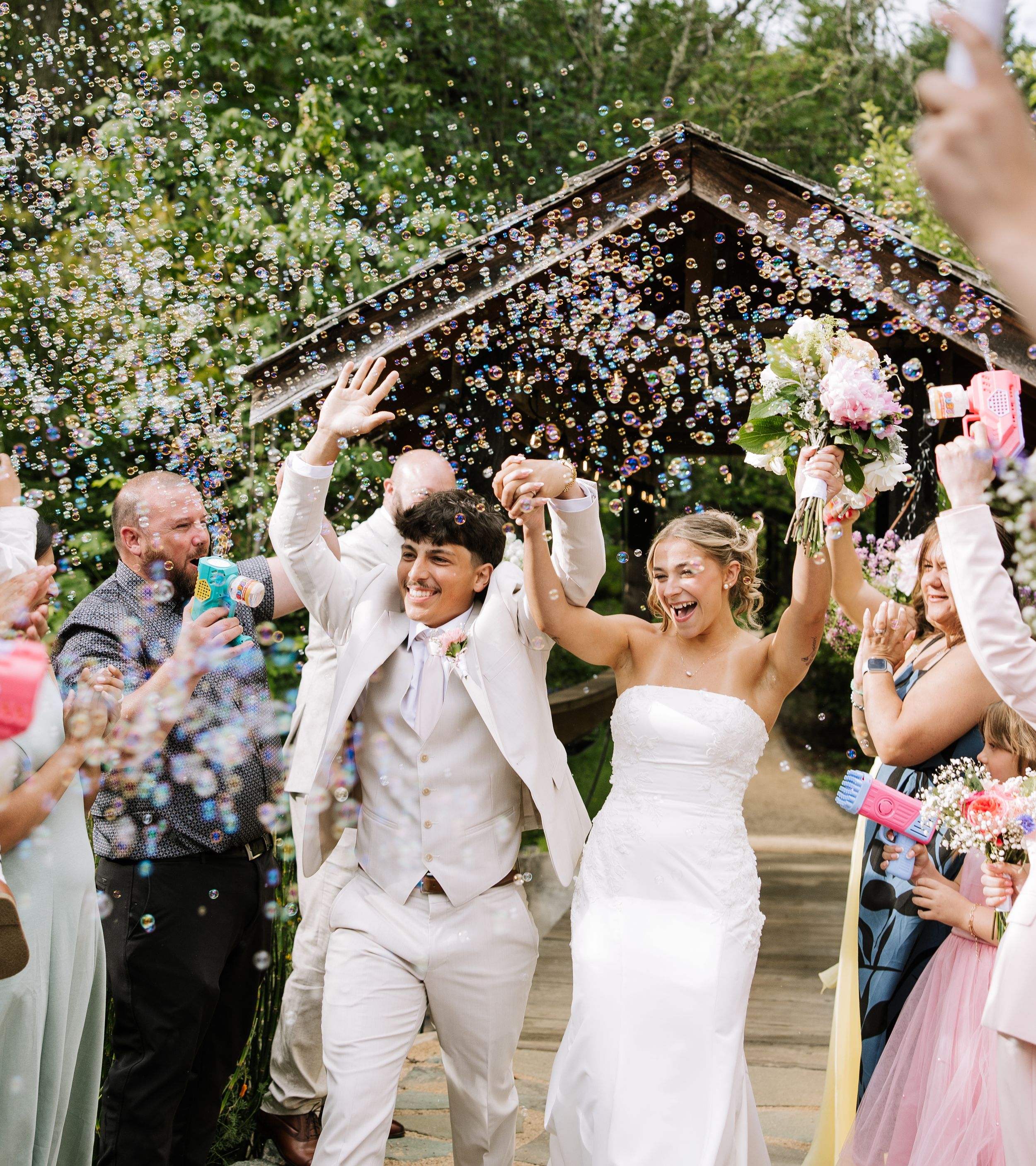 Joyful newlyweds celebrate with bubbles after outdoor wedding ceremony, surrounded by cheering guests.