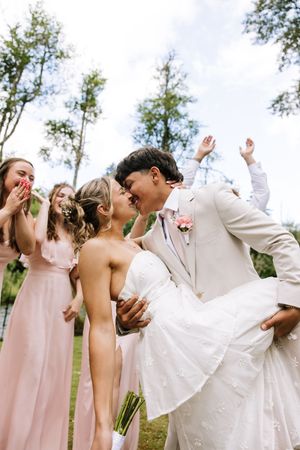Bride and groom share a romantic kiss surrounded by cheering bridesmaids in pink dresses at an outdoor wedding.