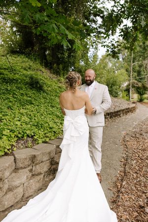 Bride in white strapless gown with bow train faces groom in tan suit on a garden path surrounded by lush greenery.