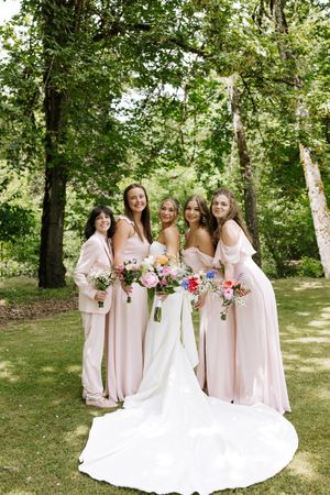 Bride in white gown poses with four bridesmaids in blush pink dresses holding colorful bouquets outdoors.