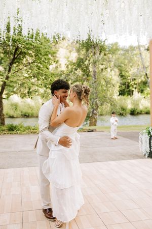 Bride and groom share first dance outdoors, surrounded by lush greenery and hanging white florals near a serene river.